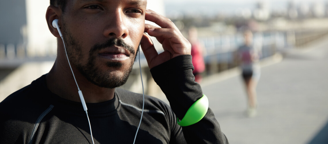 Cropped portrait of black sportsman sitting on pavement in deep thoughts, listening to motivative audiobook in his headphones, touching his head, looking confident and concentrated during workout